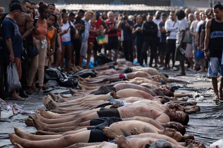 Várias pessoas deitadas lado a lado no chão, vestindo roupas íntimas, cercadas por uma multidão em ambiente externo durante o dia.