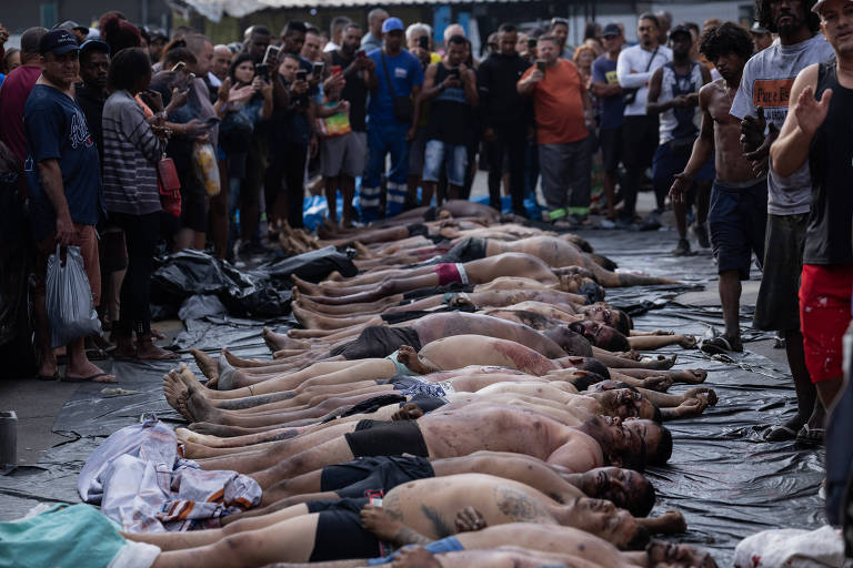 Várias pessoas deitadas lado a lado no chão, seminuas, com marcas visíveis no corpo, cercadas por uma multidão que observa em ambiente externo.