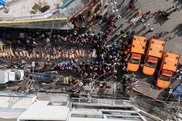Vista aérea mostra dezenas de pessoas deitadas lado a lado no meio de uma rua, cercadas por um grupo maior de manifestantes em pé. Três ambulâncias laranjas estão estacionadas próximas, e há veículos e construções ao redor. A cena ocorre em área urbana durante o dia.