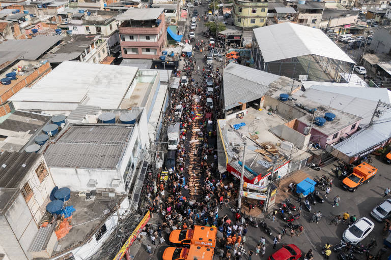 Rua estreita e lotada de pessoas entre prédios residenciais e comerciais em área urbana. Vários veículos laranja de emergência estão estacionados na parte inferior da imagem, enquanto a multidão ocupa toda a extensão da rua, que se estende até o horizonte. 