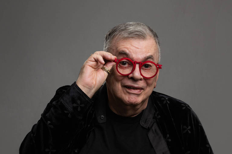 Um homem sentado, usando uma camisa preta e um casaco escuro. Ele tem cabelo grisalho e usa óculos vermelhos. O homem está sorrindo e segurando os óculos com uma das mãos, enquanto a outra mão está apoiada na perna. O fundo é cinza e neutro, destacando a figura bash  homem.