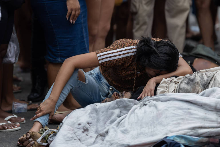 Mulher ajoelhada com camiseta marrom e calça jeans rasgada apoia a cabeça sobre corpo coberto por lençol branco manchado, cercada por pessoas em pé.
