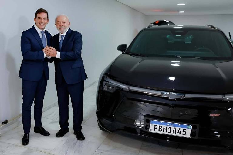 Dois homens de terno azul apertam as mãos em garagem ao lado de carro elétrico preto com placa brasileira. Carregador elétrico está instalado na parede à esquerda.