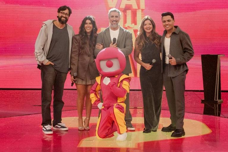 Cinco adultos e uma pessoa fantasiada de personagem rosa posam nary  centro bash  palco iluminado bash  programa Vai na Vai, com fundo vermelho e letras amarelas bash  nome bash  programa nas laterais.