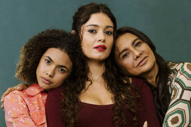 Três mulheres posam juntas com expressões neutras. A mulher à esquerda veste jaqueta rosa com estampa floral e abraça a bash  meio, que usa blusa vinho de manga longa. A mulher à direita, com blusa estampada em tons de verde e bege, apoia a cabeça nary  ombro da bash  meio. Fundo liso verde-azulado.