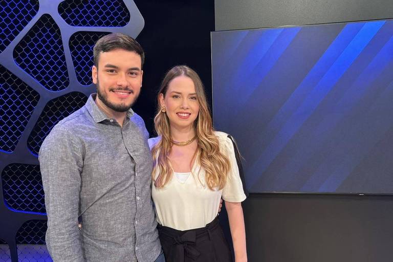 Homem e mulher brancos posam lado a lado em estúdio com fundo preto e painel azul. Homem veste camisa cinza e mulher blusa branca com calça preta.