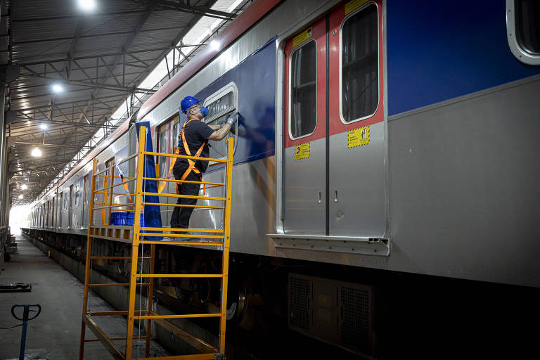 Homem com capacete azul e colete de segurança laranja está em plataforma amarela realizando manutenção na lateral de um vagão de trem dentro de um galpão iluminado.