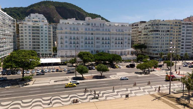 Calçadão com padrão ondulado preto e branco na orla, com ciclistas e pedestres. Avenida com carros e táxi amarelo. Ao fundo, prédios residenciais e montanha sob céu azul.