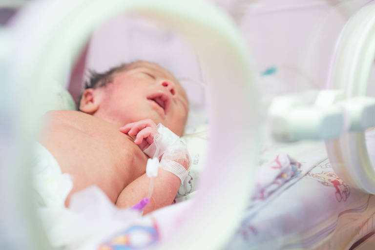 Bebê recém-nascido dorme dentro de incubadora hospitalar, com sonda intravenosa no braço esquerdo. Ambiente clínico com equipamentos médicos ao redor.