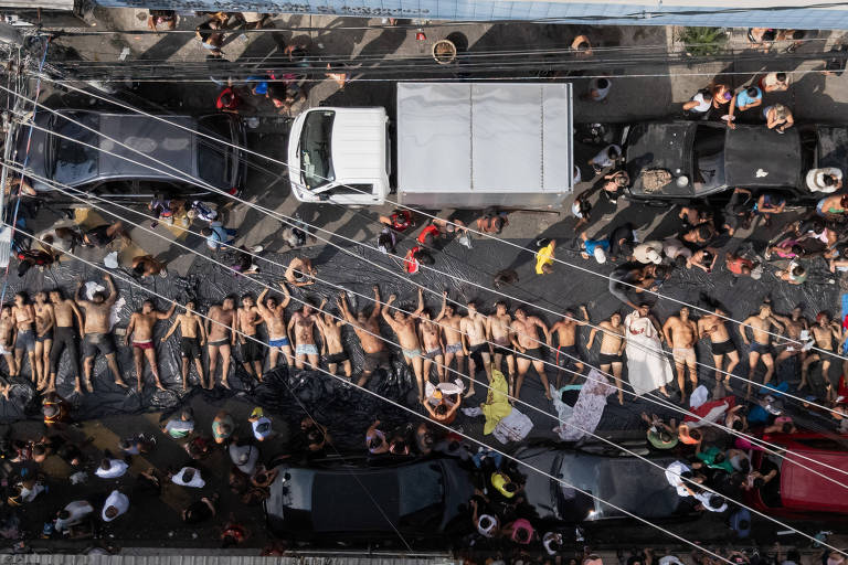 Cerca de 30 pessoas nuas deitadas lado a lado no centro de uma rua movimentada, cercadas por multidão de espectadores. Veículos estacionados e fios elétricos visíveis acima da cena.