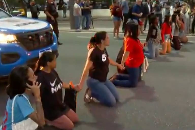 Grupo de manifestantes ajoelhados no meio de uma via urbana bloqueando o trânsito. Ao lado esquerdo, viatura policial azul e branca parada. Pessoas observam a cena na calçada ao fundo.