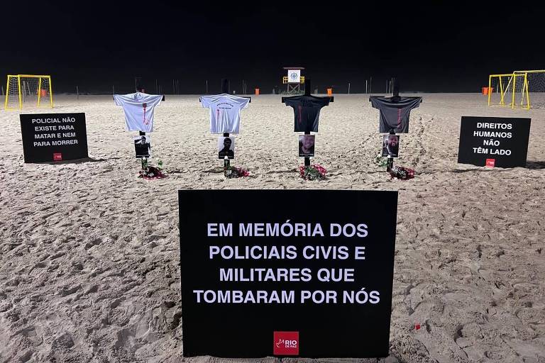 Cinco cruzes com camisas de policiais estão fincadas na areia de uma praia à noite, cercadas por placas com mensagens de memória e direitos humanos. A placa central diz: "Em memória dos policiais civis e militares que tombaram por nós". À esquerda, placa afirma: "Policial não existe para matar e sim para nos proteger". À direita, placa diz: "Direitos humanos não tem lado". Ao fundo, há dois gols de futebol de areia.