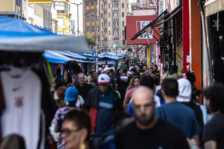 Multidão caminha por rua estreita com barracas de comércio nas laterais e placas comerciais, incluindo uma do KFC. Prédios altos cercam a área em dia claro.
