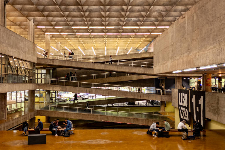 Interior da Biblioteca Central da USP com rampas em vários níveis conectando os andares. Pessoas sentadas no piso térreo conversam e estudam em grupos. Estrutura de concreto aparente e teto com iluminação linear.