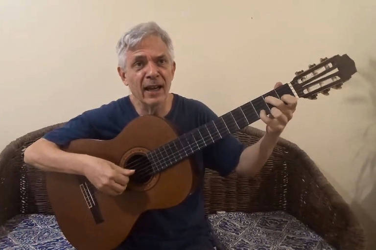 Homem de cabelos grisalhos veste camiseta azul e toca violão clássico sentado em sofá de vime com almofada estampada. Fundo é parede clara sem decoração.