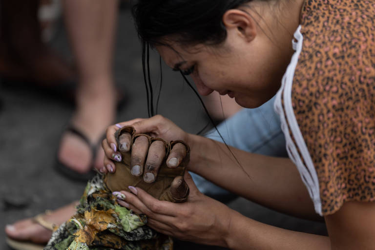 Mulher com blusa estampada segura e ajusta mão de escultura detalhada, com vegetação artificial, em ambiente externo. Pessoas ao fundo estão descalças ou de chinelos.