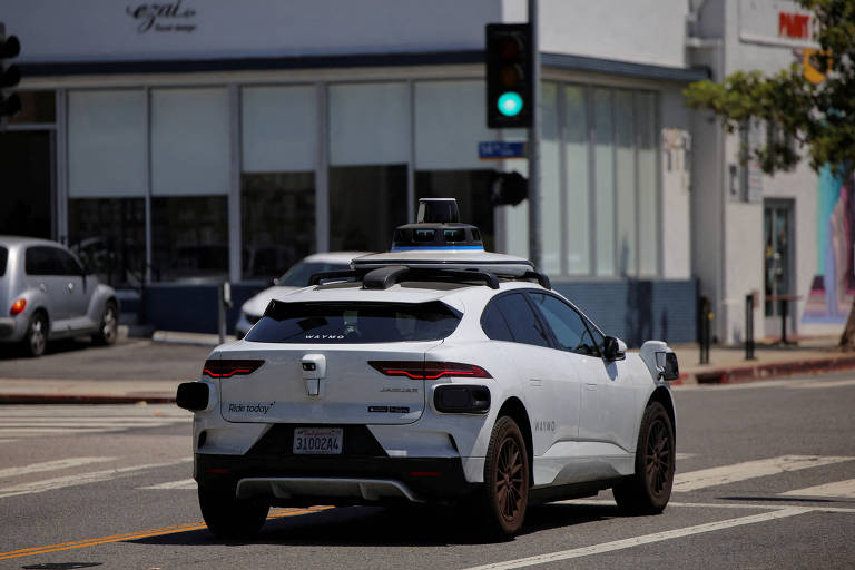 Veículo branco autônomo com sensores no teto trafega em cruzamento de rua urbana. Sinal de trânsito está verde e há prédios comerciais ao fundo.
