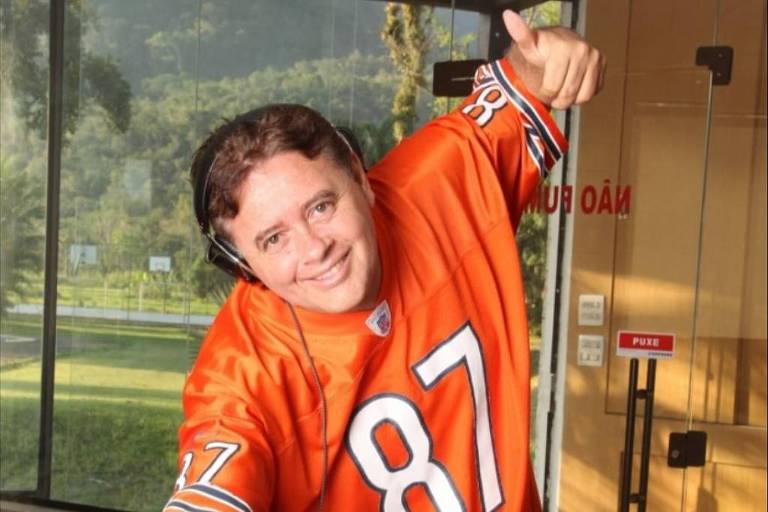 Um homem sorridente está posicionado atrás de uma mesa de DJ, usando uma camiseta laranja com o número 87. Ele está fazendo um gesto de 'joinha' com a mão direita e parece estar se divertindo. Ao fundo, há grandes janelas que mostram uma paisagem verde e uma entrada de prédio.