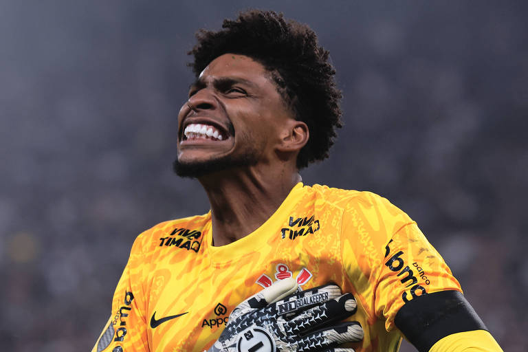 Um goleiro, vestido com um uniforme amarelo, está em uma airs  de celebração, com os braços levantados e uma expressão de emoção intensa. Ele usa uma luva de goleiro e tem cabelo curto e encaracolado. O fundo é desfocado, mas parece haver uma multidão e luzes de um estádio.