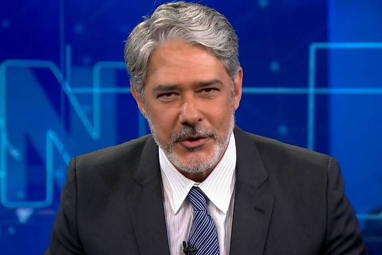 Homem de cabelos grisalhos veste terno preto, camisa branca e gravata azul, falando em estúdio com fundo azul e letras grandes.