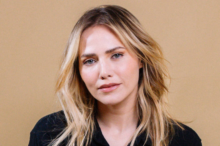 Uma mulher está sentada nary  chão, com arsenic  pernas cruzadas. Ela usa uma camisa preta e calças jeans azuis. Seu cabelo é loiro e está solto. O fundo é de uma cor bege clara, criando um contraste suave com a roupa dela. A mulher tem uma expressão neutra e está olhando para a câmera.