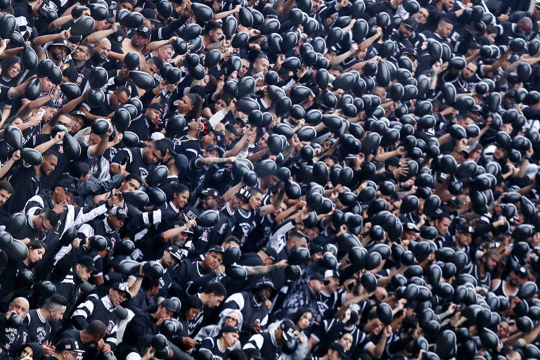 Grande grupo de torcedores vestidos com camisas pretas e bonés pretos em arquibancada de estádio, muitos com braços erguidos em comemoração ou apoio.
