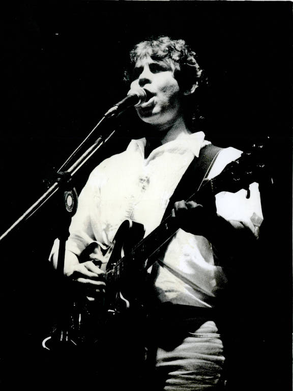 Homem jovem toca guitarra e canta em microfone dianteiro, vestido com camisa clara e calça clara, em fundo escuro.