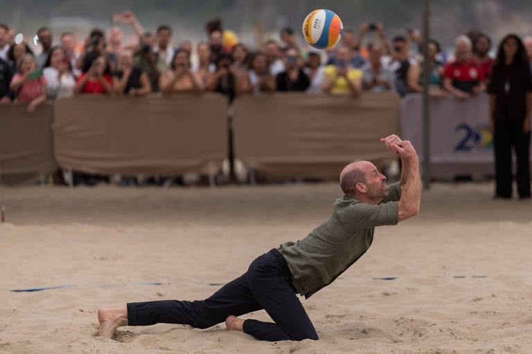 Yeşil gömlekli ve siyah pantolonlu bir adam, plaj voleybolu maçı sırasında kuma dalarak voleybol topuna ulaşmaya çalışıyor. Arka plandaki kalabalık ise oyunu dikkatle izliyor.