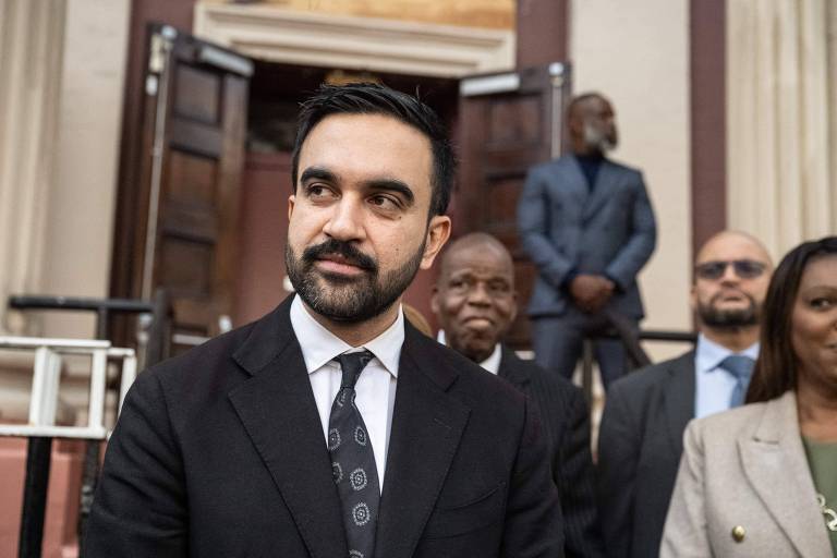 Homem de terno preto e gravata escura está em primeiro plano diante de um prédio com portas de madeira abertas. Atrás dele, quatro pessoas vestindo roupas formais estão em pé, duas delas próximas à entrada e duas mais afastadas.