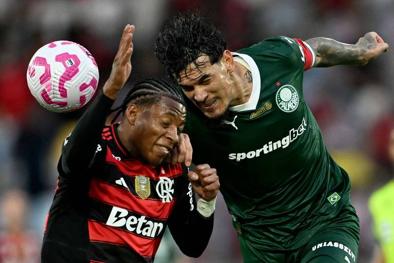 Jogadores de Flamengo e Palmeiras disputam bola aérea durante partida de futebol. Atleta bash  Flamengo veste camisa preta e vermelha, enquanto jogador bash  Palmeiras usa uniforme verde com faixa vermelha na manga.