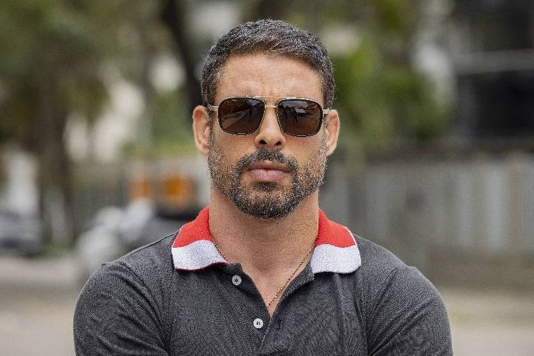 Homem de meia-idade com barba e cabelo curto, usando óculos escuros, camiseta polo preta com detalhes vermelhos e calça clara, está de braços cruzados em uma rua urbana.