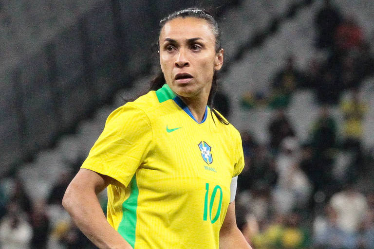 Uma jogadora de futebol está em campo, vestindo um uniforme amarelo e azul da seleção brasileira. Ela tem cabelo preso e está olhando para o lado, com uma expressão concentrada. O campo está vazio ao fundo, com algumas pessoas nas arquibancadas. O número 10 está visível em sua camisa, e ela usa meias brancas.