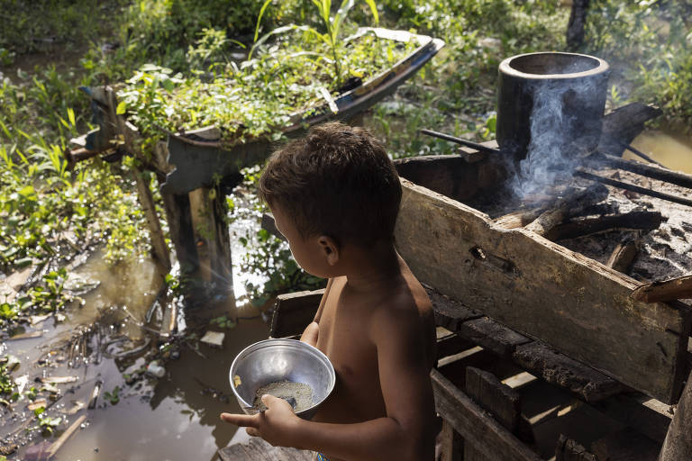 A imagem mostra uma criança sem camisa, de costas, segurando uma tigela de metal. Ao fundo, há uma fogueira com fumaça saindo e um recipiente de metal em cima dela. O ambiente é rural, com vegetação ao redor e um barco parcialmente visível. O chão está coberto por água e plantas.