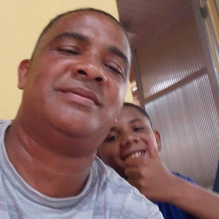 In primo piano c'è un uomo con una maglietta a righe grigie e bianche, che sorride leggermente. Dietro di lui, un ragazzo con una maglietta blu sorride e fa un cenno di saluto con la mano destra. Ambientazione interna con un muro giallo e una porta di metallo sullo sfondo.