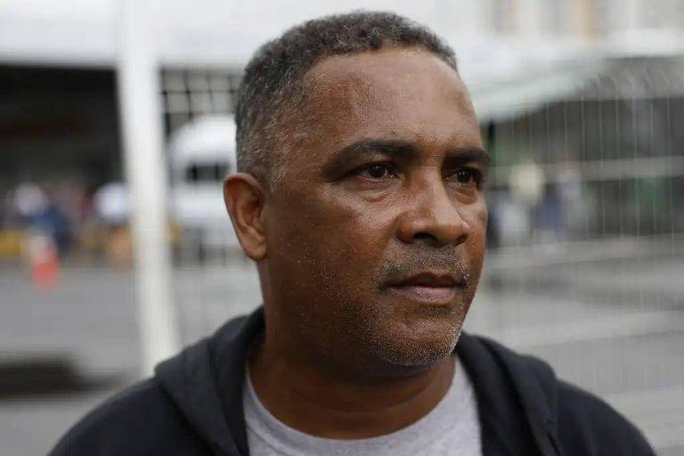 Un uomo adulto con i capelli corti e la barba incolta, che indossa una maglietta grigia e una giacca nera, guarda di lato in un ambiente esterno con una struttura metallica sfocata sullo sfondo.