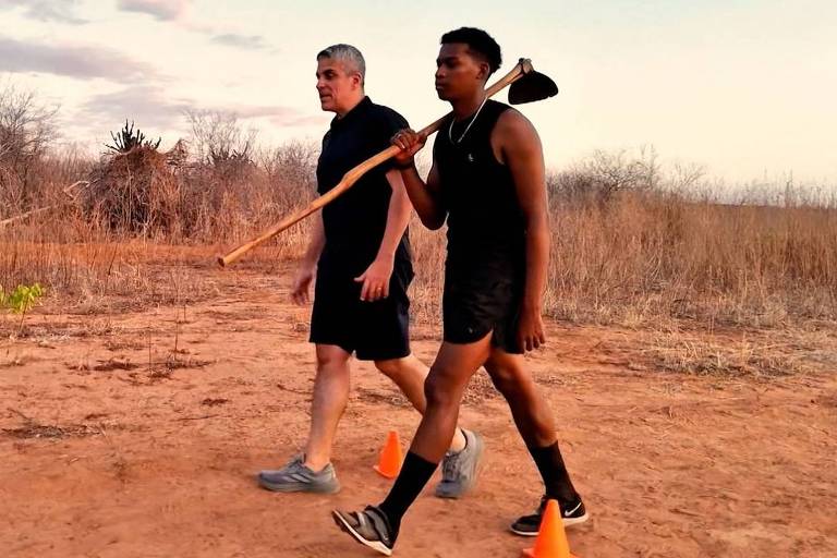 Dois homens caminham lado a lado em terreno seco e árido ao entardecer. Um deles carrega uma enxada apoiada nary  ombro direito. Vegetação rala e céu com nuvens ao fundo.