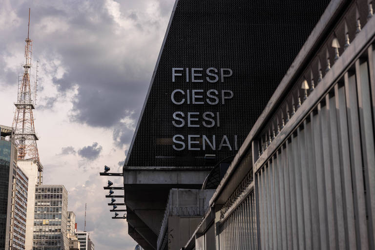 Prédio da FIESP com as siglas FIESP, CIESP, SESI e SENAI visíveis na fachada escura. Ao fundo, prédios altos e uma torre de transmissão sob céu nublado.