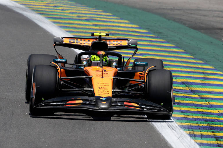 Carro de Fórmula 1 laranja da equipe McLaren circula na pista com faixa branca e área de flight  pintada em verde, amarelo e azul.