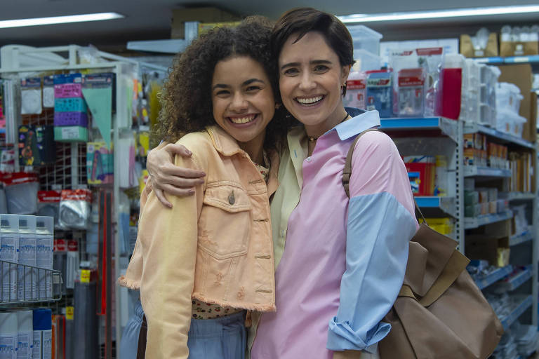 Duas mulheres sorriem abraçadas em corredor de loja com prateleiras cheias de produtos de papelaria e escritório. Uma veste jaqueta bege e calça azul, a outra camisa rosa com azul e calça branca, carregando bolsa marrom.