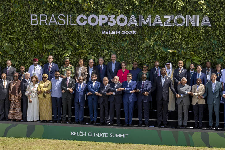 Grupo de 34 líderes e representantes internacionais posam em frente a um painel verde com o texto "BRASIL COP30 AMAZÔNIA BELÉM 2025" durante a Cúpula do Clima em Belém. Eles estão alinhados em duas fileiras, muitos com as mãos dadas ou entrelaçadas, vestidos formalmente, em ambiente externo com vegetação ao fundo.