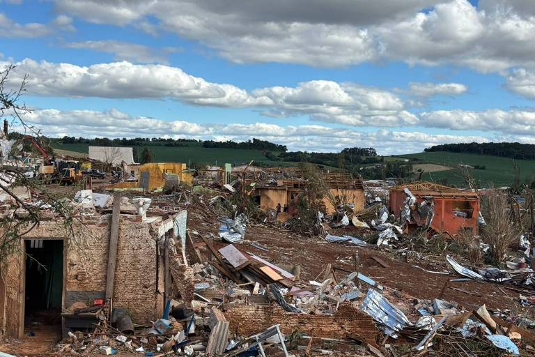 Kırsal bir alanda birkaç tuğla ev ve ahşap yapı yıkılmış durumda. Yerlere moloz, metal çatı kaplama levhaları ve molozlar dağılmış durumda. Arka planda parçalı bulutlu gökyüzü ve yeşil tepeler görülüyor.
