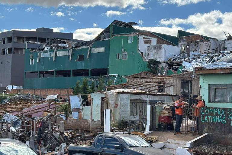 Çatısı ve duvarlarının bir kısmı yıkılmış iki katlı yeşil bir bina. Ön planda sade evler ve etrafa saçılmış molozlar. Turuncu yelekli iki kişi bir kapının yanında konuşuyor. Sokakta siyah bir araba park etmiş. Arka planda parçalı bulutlu gökyüzü.