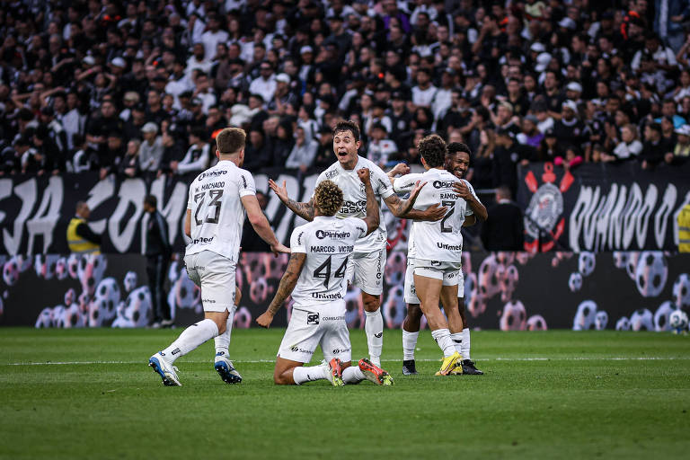 Cinco jogadores do Corinthians vestindo uniforme branco comemoram gol no campo. Um está ajoelhado, outros abraçados e um com expressão de alegria. Torcida lotada ao fundo com faixas e bandeiras em preto e branco.