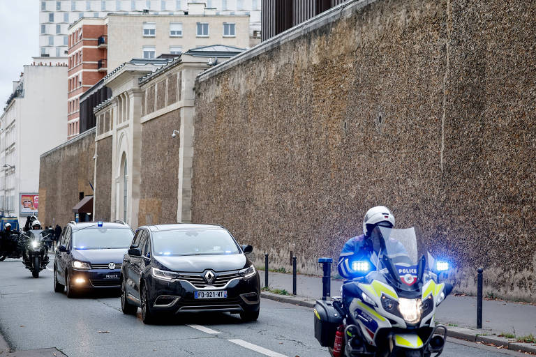 Motocicleta da polícia com luzes azuis ligadas lidera uma caravana de veículos em uma rua ao lado de um muro alto de pedra. Dois carros seguem atrás da moto, com prédios residenciais ao fundo.