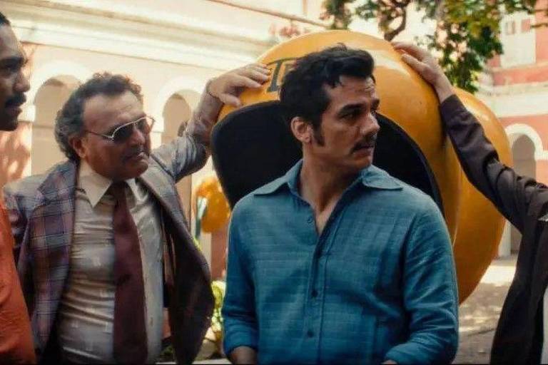 Quatro homens estão reunidos em um espaço aberto com arquitetura colonial ao fundo. Dois deles tocam uma grande bola amarela atrás do homem de camisa azul. Eles vestem roupas dos anos 1970, incluindo camisa xadrez, gravata e camisas de manga comprida.