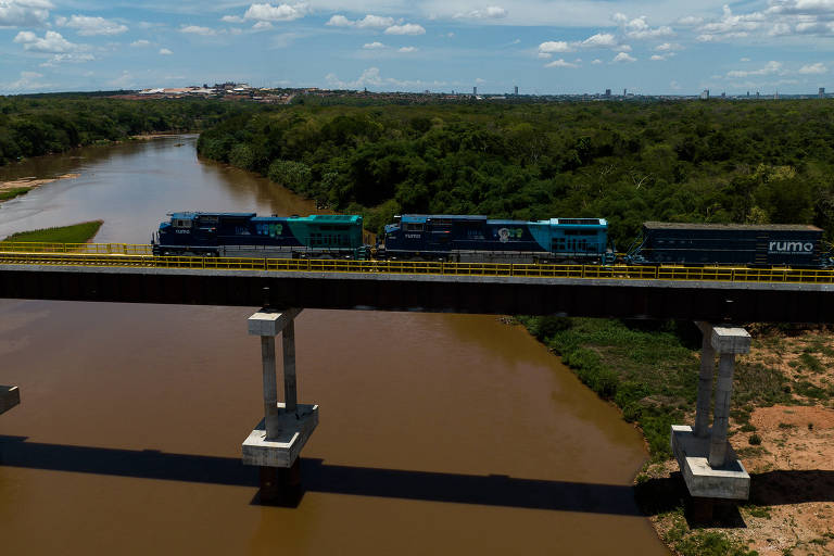 Trem de carga com locomotivas azul e verde atravessa ponte elevada sobre rio de águas barrentas. Vegetação densa e verde cobre as margens do rio sob céu parcialmente nublado.