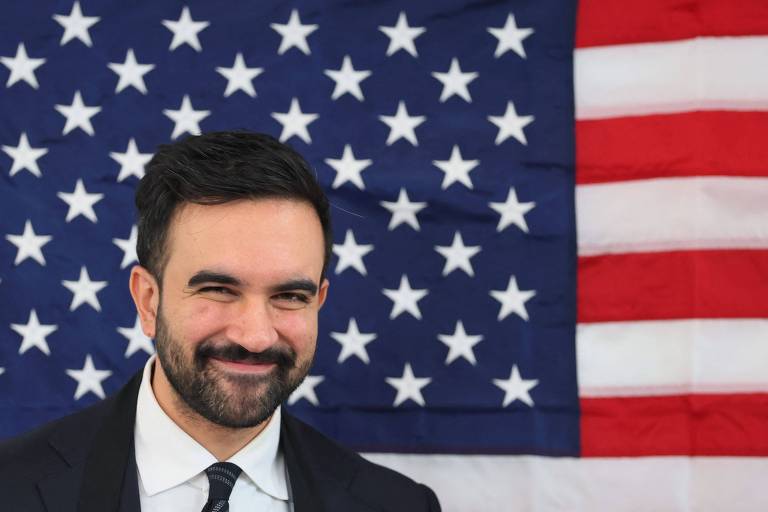 Homem de barba e cabelo escuro veste terno preto, camisa branca e gravata preta, posando com leve sorriso à frente da bandeira dos Estados Unidos, que exibe estrelas brancas sobre fundo azul e listras vermelhas e brancas.