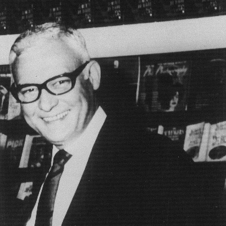 Homem de cabelos grisalhos, usando óculos escuros, terno escuro, camisa branca e gravata, sorri enquanto está em ambiente interno com prateleiras de livros ao fundo.