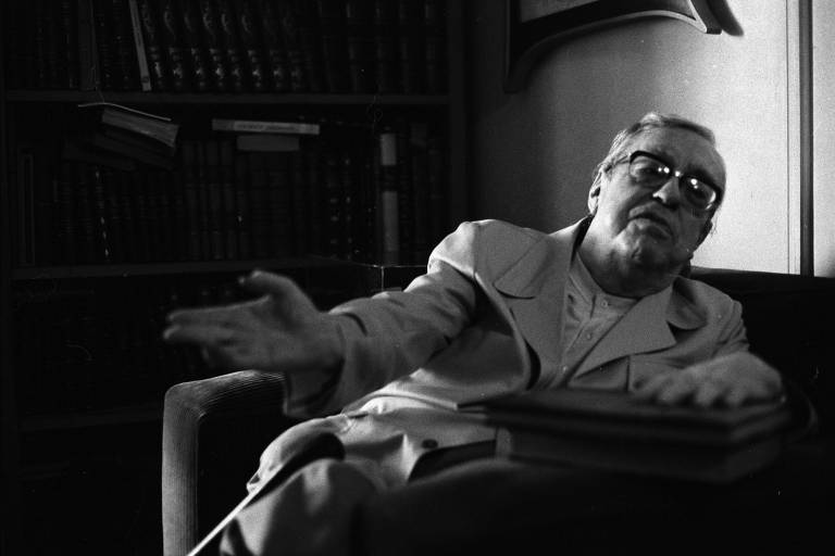 Homem idoso com óculos escuros e casaco claro está sentado em poltrona, estendendo a mão direita à frente. Atrás dele, estante cheia de livros em ambiente interno.