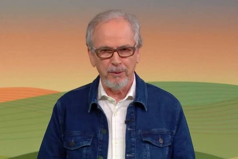 Homem idoso com cabelo grisalho e barba curta usa óculos e jaqueta jeans azul sobre camisa branca. Fundo apresenta tons degradê de laranja, verde e amarelo.